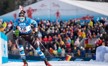 Ein Biathlet in blauem Wettkampfanzug läuft mit Skiern und Stöcken vor einer jubelnden Menge während eines Wettbewerbs.