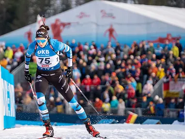 Ein Biathlet in blauem Wettkampfanzug läuft mit Skiern und Stöcken vor einer jubelnden Menge während eines Wettbewerbs.