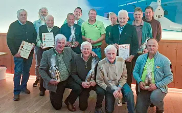 Eine Gruppe älterer Männer hält Urkunden und Flaschen, posiert lächelnd in einem Raum mit Holzboden. Im Hintergrund befindet sich ein Modell einer Kirche.