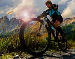 Ein Radfahrer in sportlicher Ausrüstung fährt bei Sonnenuntergang auf einem Schotterweg durch eine malerische Berglandschaft, umgeben von hohen Gipfeln und Wolken.