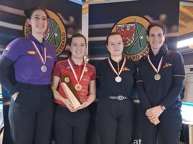 Vier Frauen mit Medaillen und Pokal stehen vor einem Banner, das mit Billard-Logos verziert ist. Sie tragen Sportkleidung und lächeln in die Kamera.