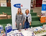 Zwei Menschen stehen an einem Messestand mit AMS-Werbung und BIZ-Material, lächeln in die Kamera. Der Tisch ist mit Broschüren und Giveaways bedeckt.