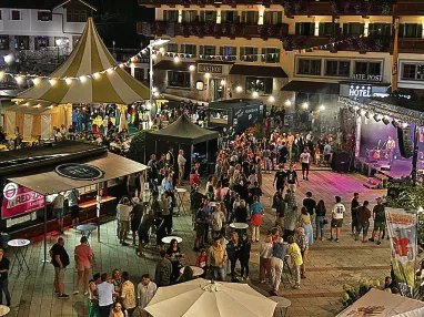Ein belebter Platz bei Nacht mit Ständen, einem Pavillon, vielen Menschen und einer beleuchteten Bühne, vor einem Gebäude mit hell erleuchteten Fenstern.