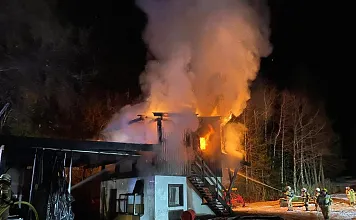 Nachtszene eines brennenden Gebäudes mit Feuerwehrleuten, die die Flammen bekämpfen. Dichtes Rauch steigt auf, während Lichter die Szene beleuchten.