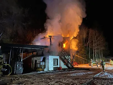 Nachtszene eines brennenden Gebäudes mit Feuerwehrleuten, die die Flammen bekämpfen. Dichtes Rauch steigt auf, während Lichter die Szene beleuchten.