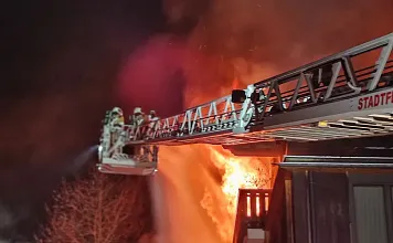 Feuerwehrleute bekämpfen einen nächtlichen Brand von einer Leiter aus, während Flammen aus einem Gebäude schlagen.