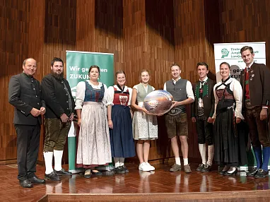 Eine Gruppe von Männern und Frauen in traditioneller bayerischer Tracht steht auf einer Bühne mit Holzwänden und lächelt in die Kamera.
