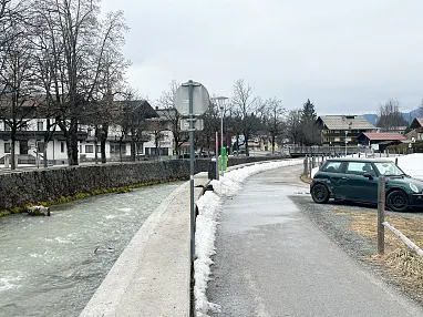 Verschneiter Weg entlang eines Flusses, mit parkendem Auto rechts. Im Hintergrund sind Häuser, Bäume und ein bewölkter Himmel zu sehen.