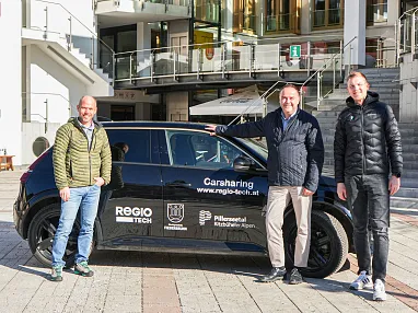 Drei Männer stehen vor einem schwarzen Auto mit Carsharing-Werbung auf einem Platz, umgeben von modernen Gebäuden und Treppen.
