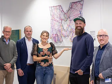 Fünf Personen stehen lächelnd vor einer farbenfrohen, fadenbespannten Kunstinstallation an der Wand in einem modernen Raum.