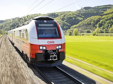 Moderner ÖBB-Zug auf Schienen, umgeben von grüner Landschaft und Hügeln im Hintergrund, bei sonnigem Wetter fahrend. Kein erkennbares Ziel oder Passagiere sichtbar.