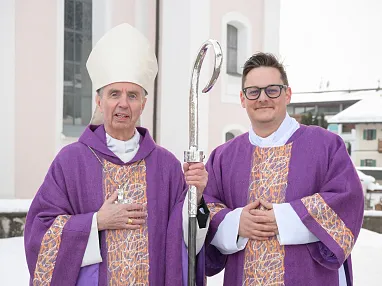 Zwei Geistliche in lila Gewändern und Bischofsmütze stehen vor einer Kirche im Schnee. Der eine hält einen Bischofsstab und beide lächeln in die Kamera.