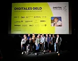 Gruppe von Menschen posieren vor einer Leinwand, auf der Veranstaltung "Digitales Geld" angekündigt wird. Sponsorenlogos und ein Bild sind sichtbar.