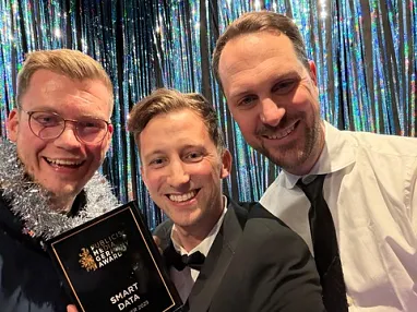 Drei fröhliche Männer in Anzügen posieren vor einem glitzernden Hintergrund, einer hält eine Trophäe mit dem Schriftzug "Award". Feierliche Stimmung ist spürbar.