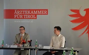 Zwei Männer sitzen an einem Tisch vor einem Banner der Ärztekammer Tirol. Vor ihnen stehen Mikrofone und eine Wasserflasche.