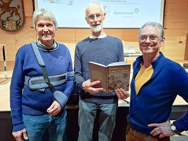 Drei Männer stehen in einem Raum, einer hält ein Buch in der Hand. Sie lächeln und tragen Freizeitkleidung. Im Hintergrund ist ein Logo an der Holzwand.