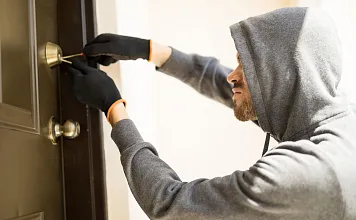 Person mit Kapuzenpullover und Handschuhen versucht, eine Tür mit einem Werkzeug zu öffnen. Die Szene wirkt angespannt und geheimnisvoll.