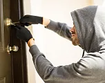 Person mit Kapuzenpullover und Handschuhen versucht, eine Tür mit einem Werkzeug zu öffnen. Die Szene wirkt angespannt und geheimnisvoll.