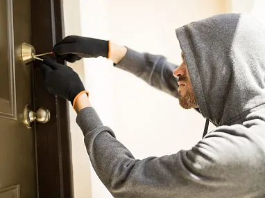 Person mit Kapuzenpullover und Handschuhen versucht, eine Tür mit einem Werkzeug zu öffnen. Die Szene wirkt angespannt und geheimnisvoll.