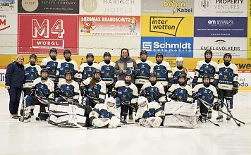 Eishockeyteam in blauen Trikots posiert für ein Gruppenfoto auf dem Eis. Trainer steht in der Mitte, mehrere Logos an der Stadionwand im Hintergrund.