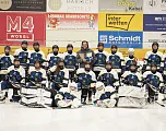 Eishockeyteam in blauen Trikots posiert für ein Gruppenfoto auf dem Eis. Trainer steht in der Mitte, mehrere Logos an der Stadionwand im Hintergrund.