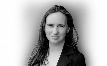 Schwarz-Weiß-Porträt einer Person mit langen Haaren, die leicht lächelt. Sie trägt eine Jacke über einem hellen Oberteil vor neutralem Hintergrund.