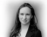 Schwarz-Weiß-Porträt einer Person mit langen Haaren, die leicht lächelt. Sie trägt eine Jacke über einem hellen Oberteil vor neutralem Hintergrund.