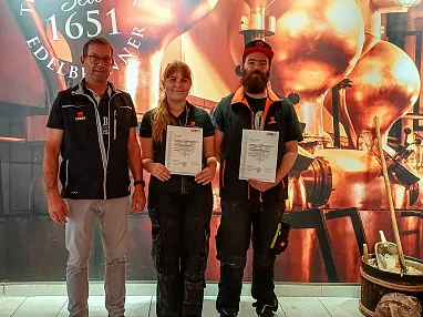 Drei Personen posieren vor einer Kupferdestillerie-Kulisse. Zwei von ihnen halten Urkunden, die sie für eine besondere Leistung erhalten haben.