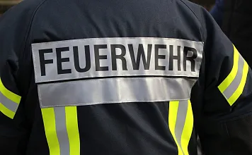 Das Bild zeigt die Rückseite einer Feuerwehrjacke mit reflektierenden Streifen und der Aufschrift "FEUERWEHR" in großen Buchstaben auf einem grauen Streifen.