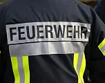 Das Bild zeigt die Rückseite einer Feuerwehrjacke mit reflektierenden Streifen und der Aufschrift "FEUERWEHR" in großen Buchstaben auf einem grauen Streifen.