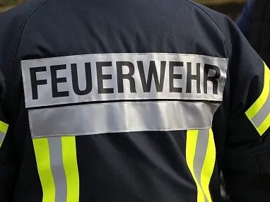 Das Bild zeigt die Rückseite einer Feuerwehrjacke mit reflektierenden Streifen und der Aufschrift "FEUERWEHR" in großen Buchstaben auf einem grauen Streifen.