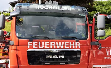 Rotes Feuerwehrauto der Marke MAN mit der Aufschrift "Hochfilzen" und "Feuerwehr" auf der Front. Teilweise verdeckte Fahrerkabine, im Freien abgestellt.