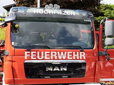 Rotes Feuerwehrauto der Marke MAN mit der Aufschrift "Hochfilzen" und "Feuerwehr" auf der Front. Teilweise verdeckte Fahrerkabine, im Freien abgestellt.