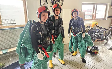 Drei Personen in Schutzanzügen und Helmen stehen in einer Garage mit Fahrrädern und Ausrüstung. Sie tragen auch gelbe Gummistiefel und Handschuhe.