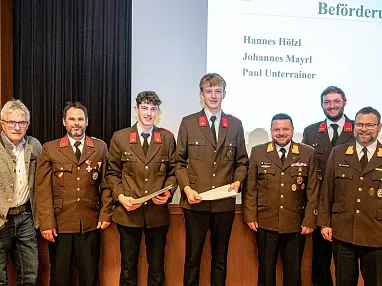 Eine Gruppe uniformierter Feuerwehrmänner posiert auf einer Bühne vor einer Leinwand während einer Veranstaltung in einem Saal.