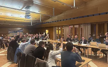 Menschen sitzen an Tischen in einem großen Raum und heben die Hände, möglicherweise bei einer Abstimmung. Der Raum hat eine Holzdecke und große Fenster.