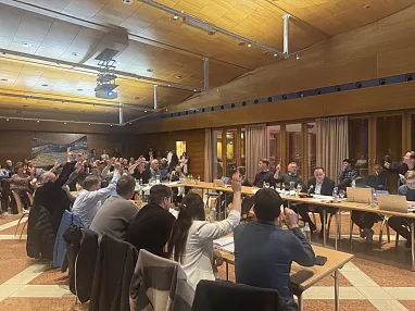 Menschen sitzen an Tischen in einem großen Raum und heben die Hände, möglicherweise bei einer Abstimmung. Der Raum hat eine Holzdecke und große Fenster.