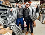 Zwei Personen stehen in einem Lager voller Bau- oder Installationsmaterialien und halten große Kabel oder Schläuche. Sie lächeln in die Kamera.