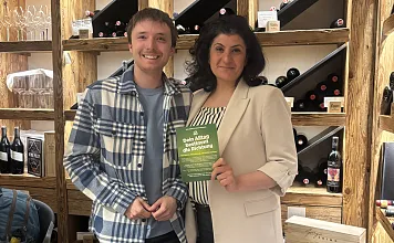 Ein Mann und eine Frau stehen nebeneinander und halten ein grünes Buch. Im Hintergrund sind Regale mit Weinflaschen und Holzkisten zu sehen.