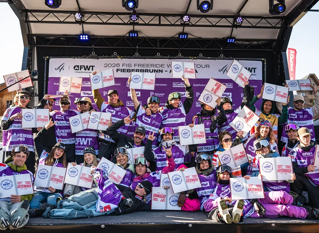 Eine Gruppe von Wintersportlern posiert auf einer Bühne mit Zertifikaten, umgeben von Bannern und Scheinwerfern. Sie tragen Winterkleidung und wirken jubelnd.