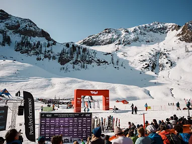 Menschen versammeln sich auf schneebedecktem Berg vor einem roten Torbogen bei einem Wintersport-Event mit blauen Himmel und verschneiten Alpen im Hintergrund.