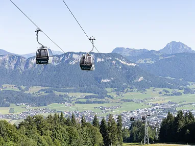 Zwei Gondeln schweben über eine grüne Alpinlandschaft mit Bergen und einem Tal, das von Bäumen umgeben ist. Der Himmel ist klar und blau.