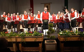 Eine traditionelle Blaskapelle in Trachten auf einer Bühne, umgeben von Blumengestecken, posiert nach einem Auftritt für ein Gruppenfoto.