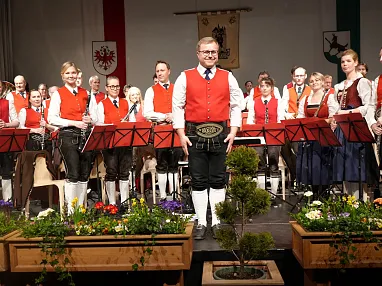 Eine traditionelle Blaskapelle in Trachten auf einer Bühne, umgeben von Blumengestecken, posiert nach einem Auftritt für ein Gruppenfoto.