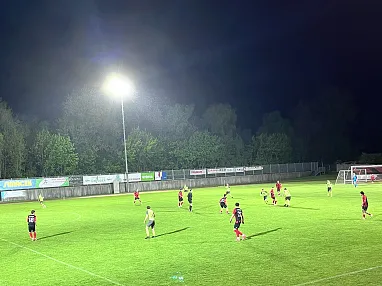 Ein nächtliches Fußballspiel auf einem beleuchteten Spielfeld, umgeben von Bäumen. Spieler in roten und schwarzen Trikots befinden sich im Spiel.