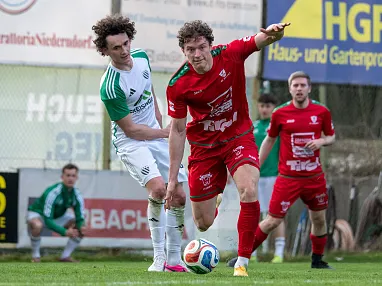 Zwei Fußballspieler im Zweikampf auf einem Spielfeld, einer in rotem Trikot, der andere in weiß-grün. Weitere Spieler und Werbetafeln im Hintergrund.