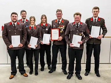 Eine Gruppe von sieben Menschen in Uniformen hält Zertifikate und eine Urkunde. Sie stehen lächelnd nebeneinander vor einer weißen Wand.