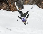 Ein Skifahrer macht einen akrobatischen Rückwärtssalto auf einer schneebedeckten Piste, umgeben von Felsen. Der Skifahrer trägt Schutzkleidung und eine Skibrille.
