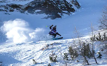 Ein Skifahrer fährt dynamisch einen verschneiten Hang hinab, umgeben von Bäumen und einer Bergkulisse unter blauem Himmel.