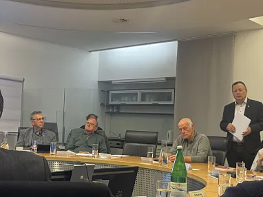 Menschen in einem Konferenzraum sitzen um einen Tisch, während eine Person vorne steht und spricht, an der Wand ein großer Bildschirm.